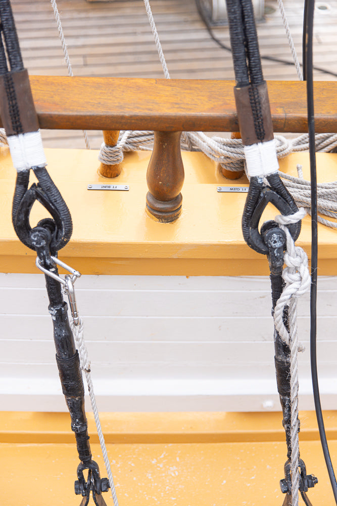 Close-up of two heavy-duty ropes attached to metal shackles on a wooden deck of a ship. The ropes are secured with white tape and lead to metal fixtures.