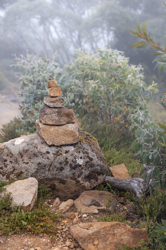A cairn, a stack of balanced stones, sits on a large rock on a misty mountain trail. Green foliage and trees are visible in the soft, hazy background.