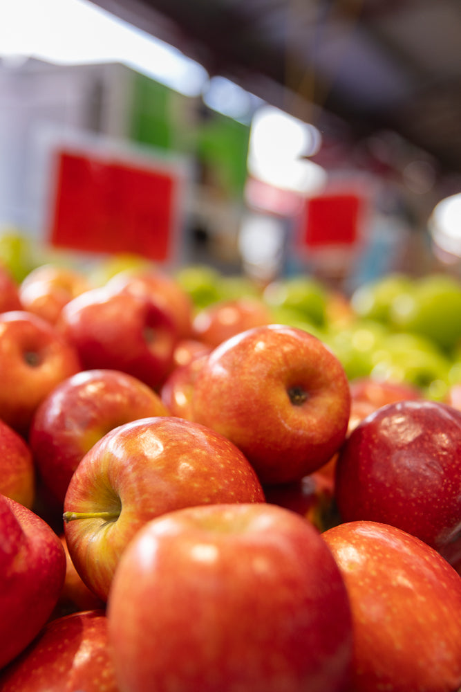 A close-up shot of a pile of red apples in a grocery store. The apples are fresh and shiny, with some green apples visible in the background. The lighting is bright, highlighting the texture and color of the fruit.