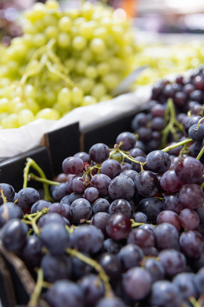A close-up shot shows two types of grapes. In the foreground, dark purple grapes are clustered together, with some stems visible. In the background, out of focus, are lighter green grapes.