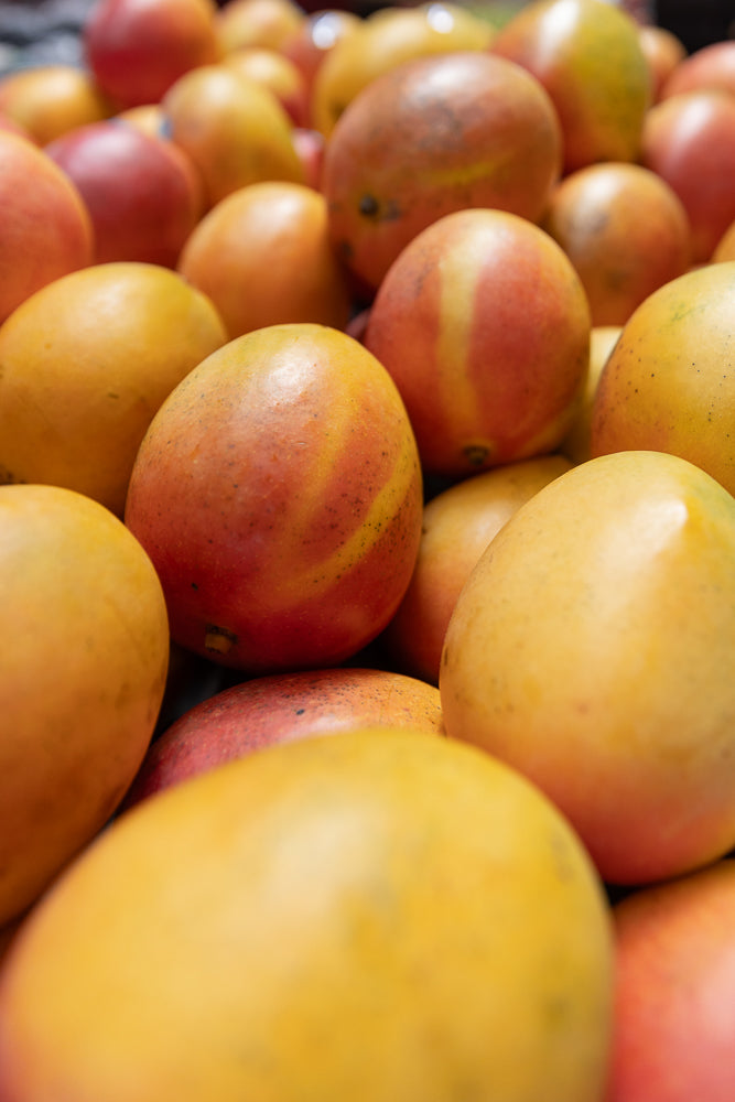 A close-up, shallow depth of field shot shows a pile of ripe mangoes. The mangoes are a mix of yellow and red, with some showing streaks of both colors. The focus is on the mangoes in the center of the frame, with the foreground and background slightly blurred.