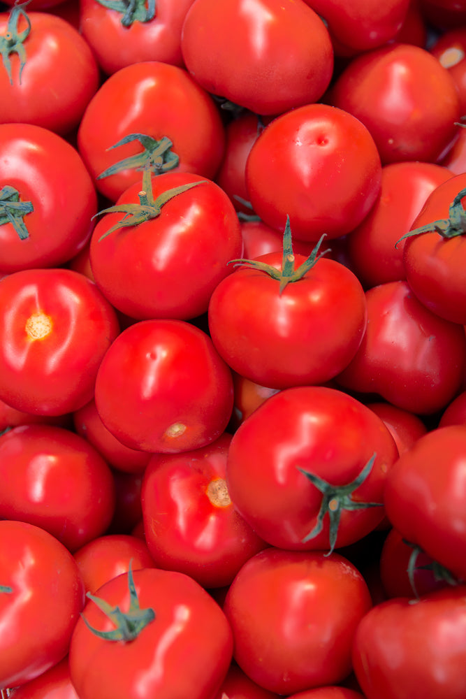 A close-up, overhead view of a pile of ripe, red tomatoes. The tomatoes are round and smooth, with some showing green stems and leaves. The lighting highlights the glossy skin of the tomatoes.