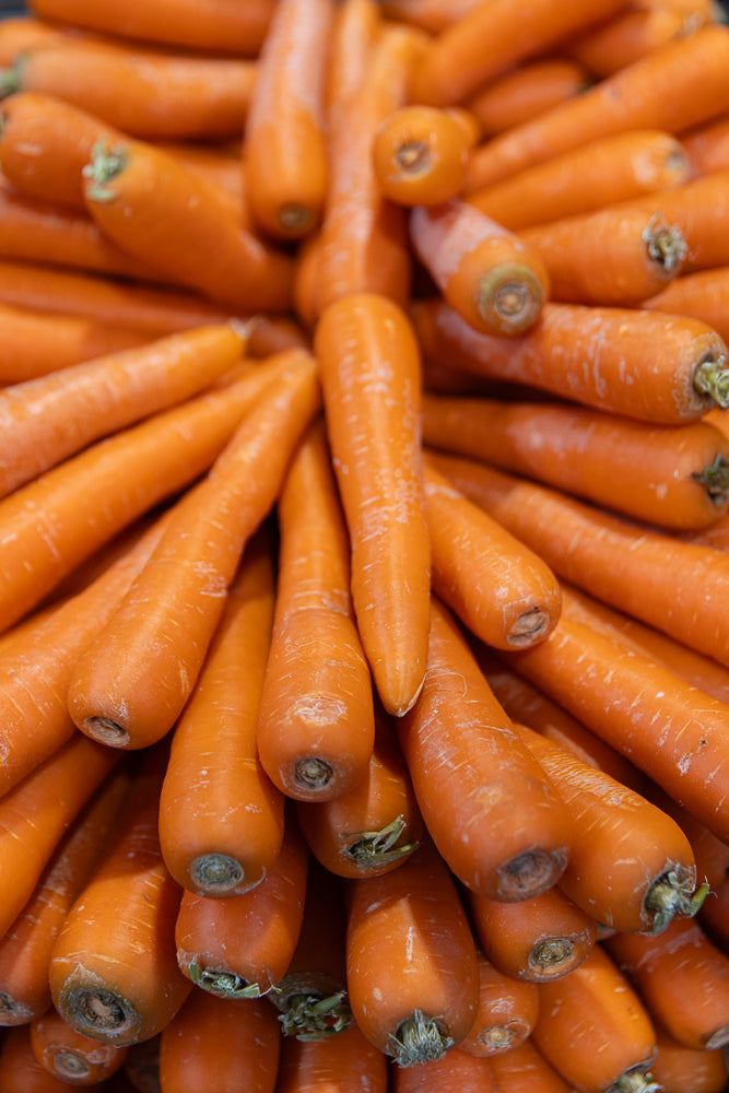 A close-up, overhead view of a large pile of fresh, bright orange carrots. The carrots are arranged in a circular pattern, with their leafy green tops pointing inwards and their tapered ends fanning outwards. Some carrots have slight imperfections or white markings on their skin.