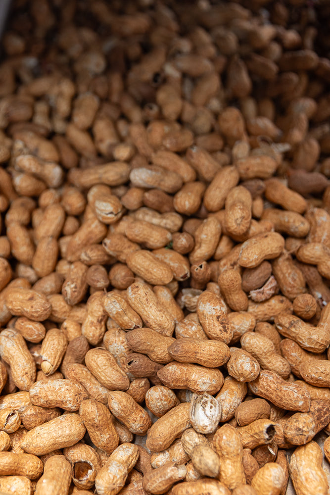 A close-up, overhead view of a large pile of unshelled peanuts. The peanuts are light brown with textured shells and are densely packed together.