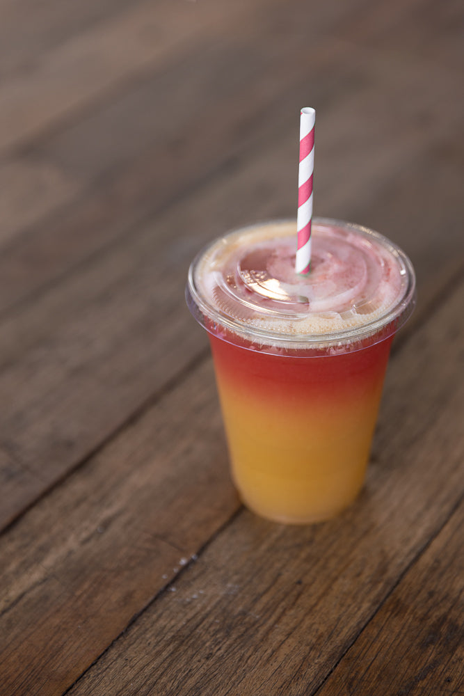 A layered drink in a clear plastic cup with a red and white striped straw. The drink has a gradient from yellow at the bottom to pink at the top, with foam on the surface.
