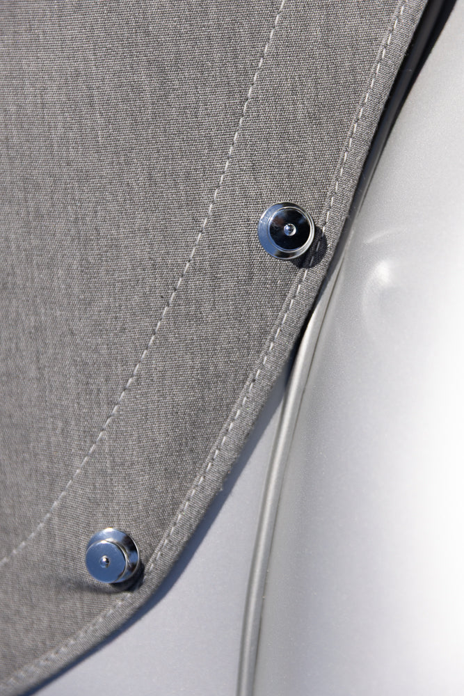 A close-up shot shows a textured gray fabric with white stitching. Two shiny silver snap fasteners are attached to the fabric, which curves over a smooth, reflective silver surface.