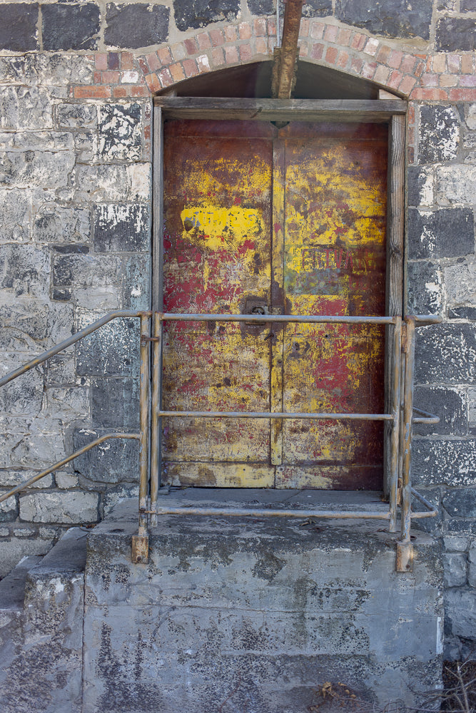 A weathered, double metal door with peeling yellow and red paint is set into a rough stone wall. A metal handrail leads up to the door, which is accessed by concrete steps.