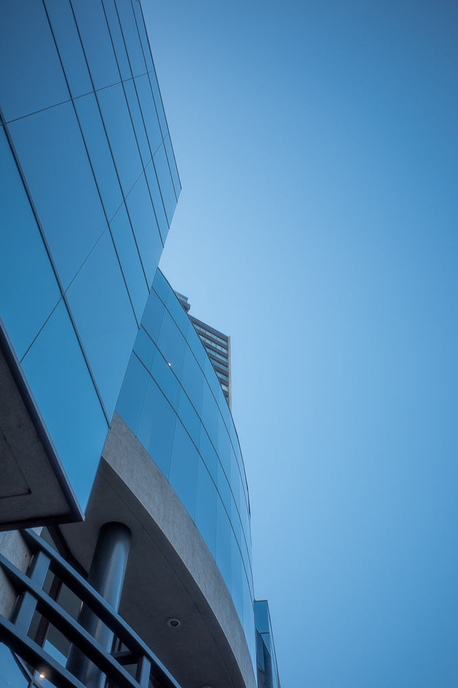 A low-angle shot of a modern building with a curved glass facade against a clear blue sky. The glass reflects the sky, creating a sleek, contemporary aesthetic.