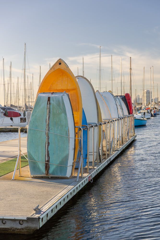 A row of colorful dinghies are stored upside down on a dock in a marina. The boats are yellow, blue, white, and red. In the background, many sailboats are moored, and a city skyline is visible in the distance.