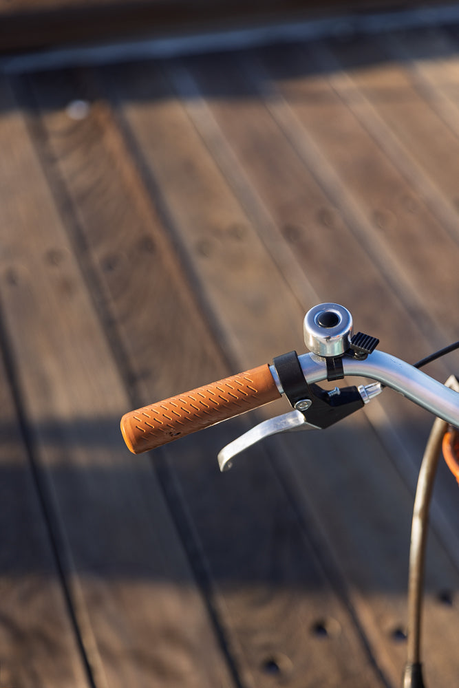 Close-up of a bicycle handlebar with a brown textured grip and a silver bell. A brake lever is visible below the grip. The background is a blurred wooden surface.