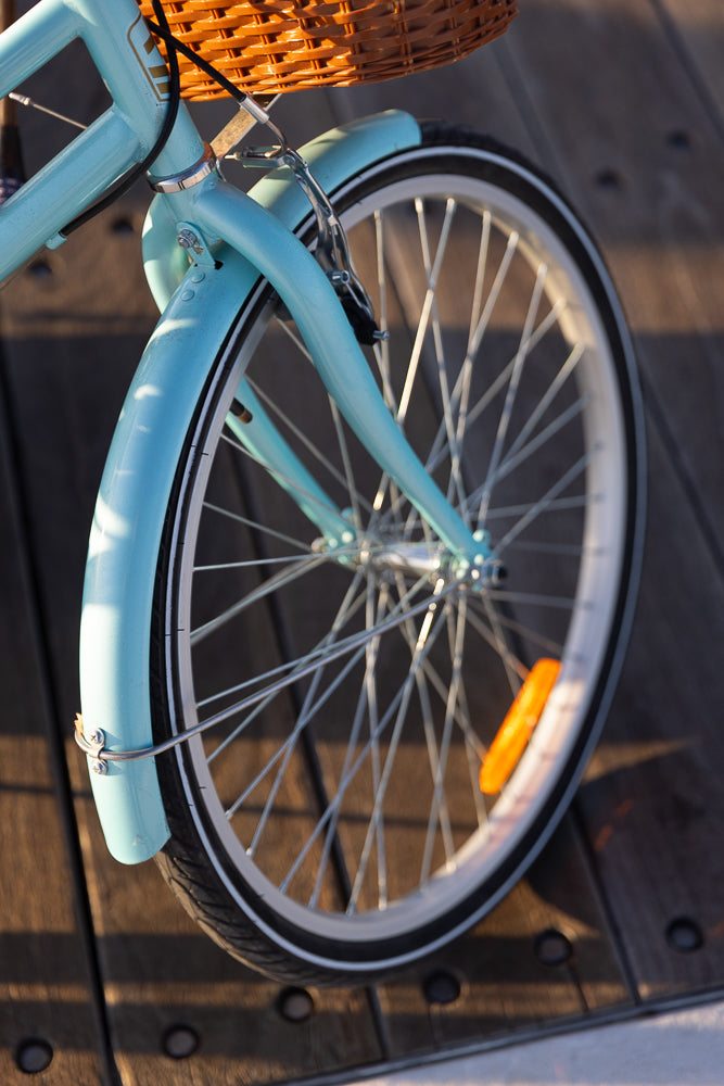 Close-up of the front wheel and fender of a light blue bicycle. The bicycle has a wicker basket attached to the handlebars and is parked on a wooden deck.