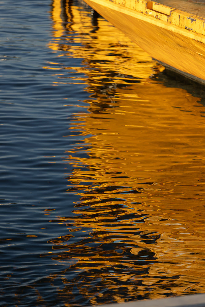 The reflection of a yellow structure on dark blue water. The water has ripples, distorting the reflection into abstract shapes and patterns.