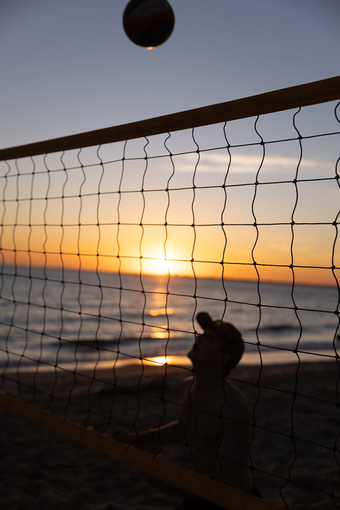 A volleyball is in mid-air against a sunset sky, with the sun setting over the ocean. A volleyball net is in the foreground, and a blurred figure of a person playing volleyball is visible behind the net.