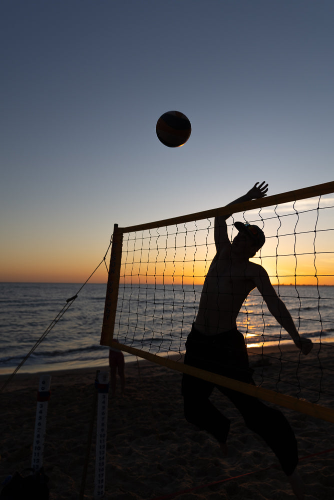 Silhouette of a beach volleyball player jumping to hit the ball over the net at sunset. The sky is a gradient of blue to orange, and the ocean is visible in the background.