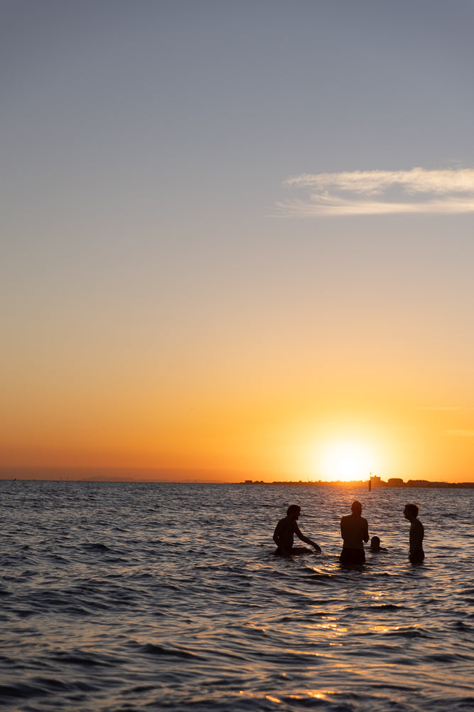 Silhouettes of four people wading in the ocean at sunset. The sun is low on the horizon, casting a warm orange glow across the sky and reflecting on the water.