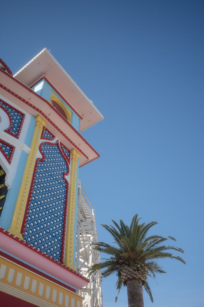 A low-angle shot of a colorful building with a roller coaster structure behind it, and a palm tree in the foreground against a clear blue sky.