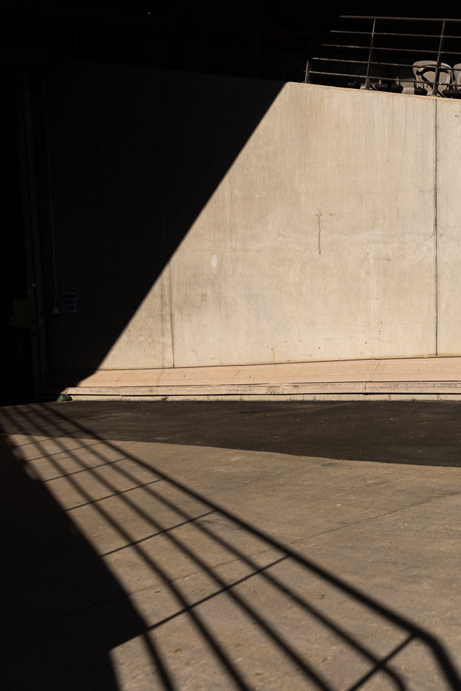 A concrete wall is partially covered by a large shadow, creating a diagonal line across the surface. Below the wall, a dark asphalt surface is crossed by the parallel shadows of a railing.