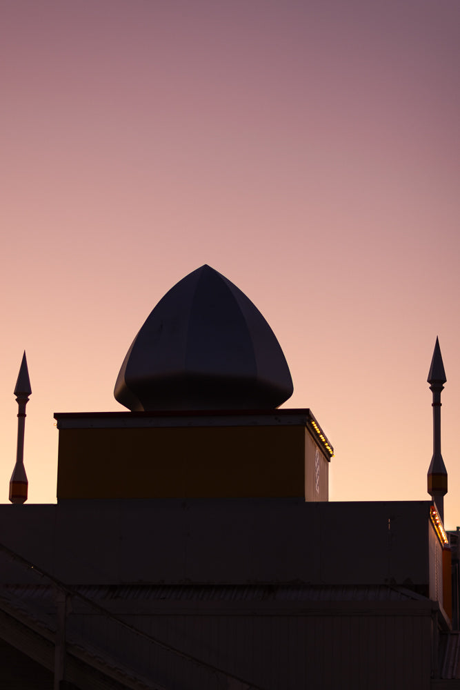 Silhouette of a building with a dome and two spires against a soft pink and purple sunset sky. The building has a yellow section with decorative lights.