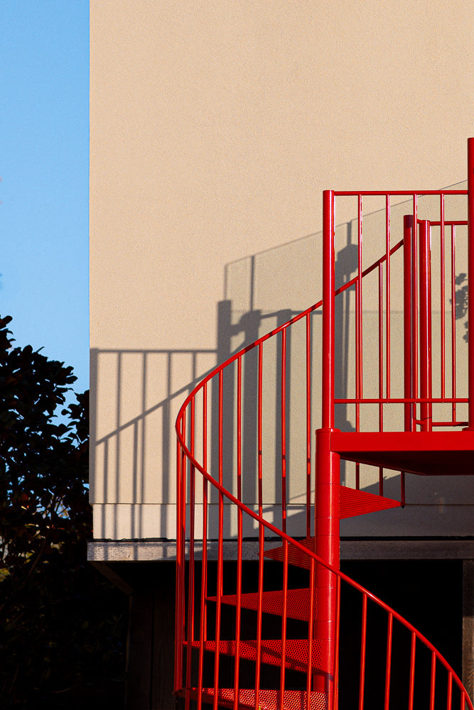 A bright red spiral staircase is shown against a beige wall and a blue sky. The staircase casts a shadow on the wall. Dark foliage is visible on the left side of the image.