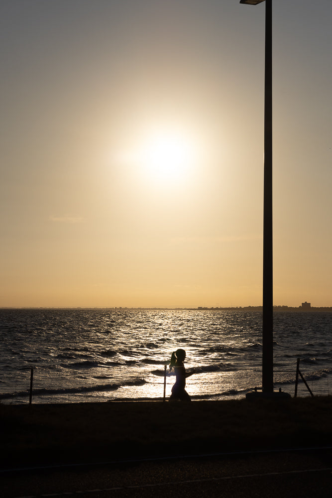 Silhouette of a person with a ponytail walking along the shore at sunset. The sun reflects off the choppy water, and a tall lamppost stands to the right.