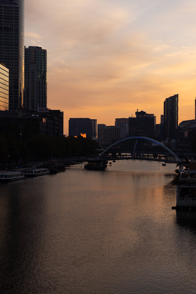 A city skyline at sunset with a river in the foreground. The sky is filled with warm orange and yellow hues, reflecting on the water. Several buildings, including tall skyscrapers and a bridge, are silhouetted against the colorful sky. Boats are visible on the river.
