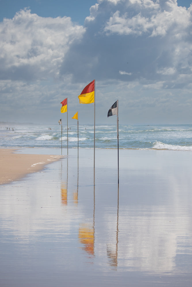A line of surf lifesaving flags, including red and yellow, yellow, and black and white checkered flags, stand on a wet sandy beach. The flags are reflected in the shallow water covering the sand. In the background, gentle waves roll onto the shore under a cloudy sky.