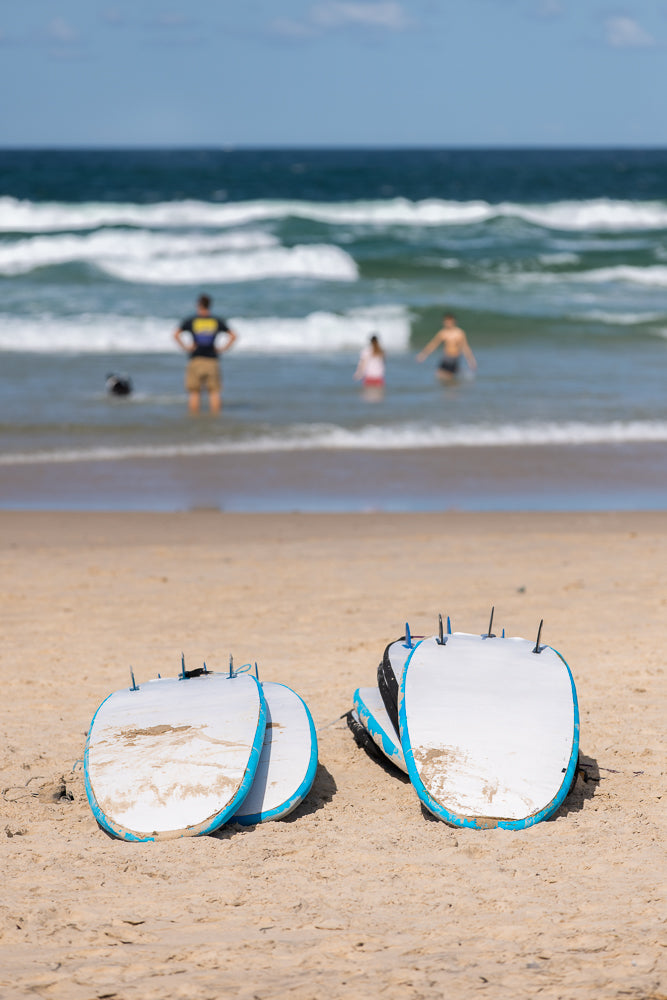 Two white surfboards with blue trim are propped up on a sandy beach. In the background, people and a dog are in the ocean with waves.