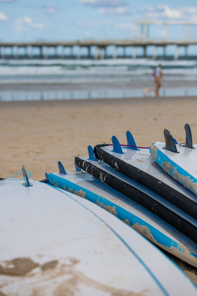 A stack of surfboards with blue and black fins rests on a sandy beach. In the background, a pier stretches into the ocean under a partly cloudy sky, and a person walks along the shoreline.