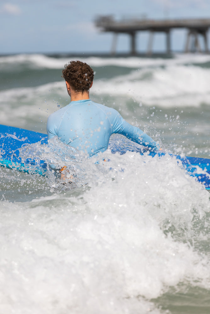A surfer in a blue rash guard paddles on a blue surfboard through choppy ocean water, with a pier visible in the blurred background.