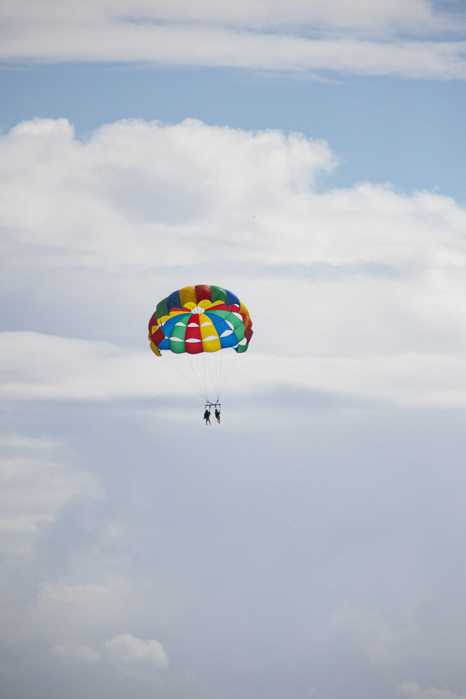 Two people parasailing high in the sky with a colorful parachute against a backdrop of white clouds and blue sky.
