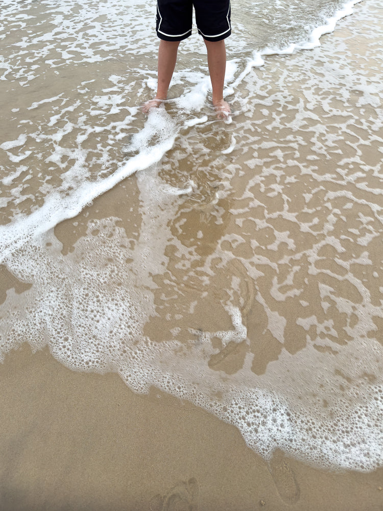 A person stands with their feet in the shallow water at the edge of the ocean. The water is clear and foamy, with small waves lapping at the sandy shore.
