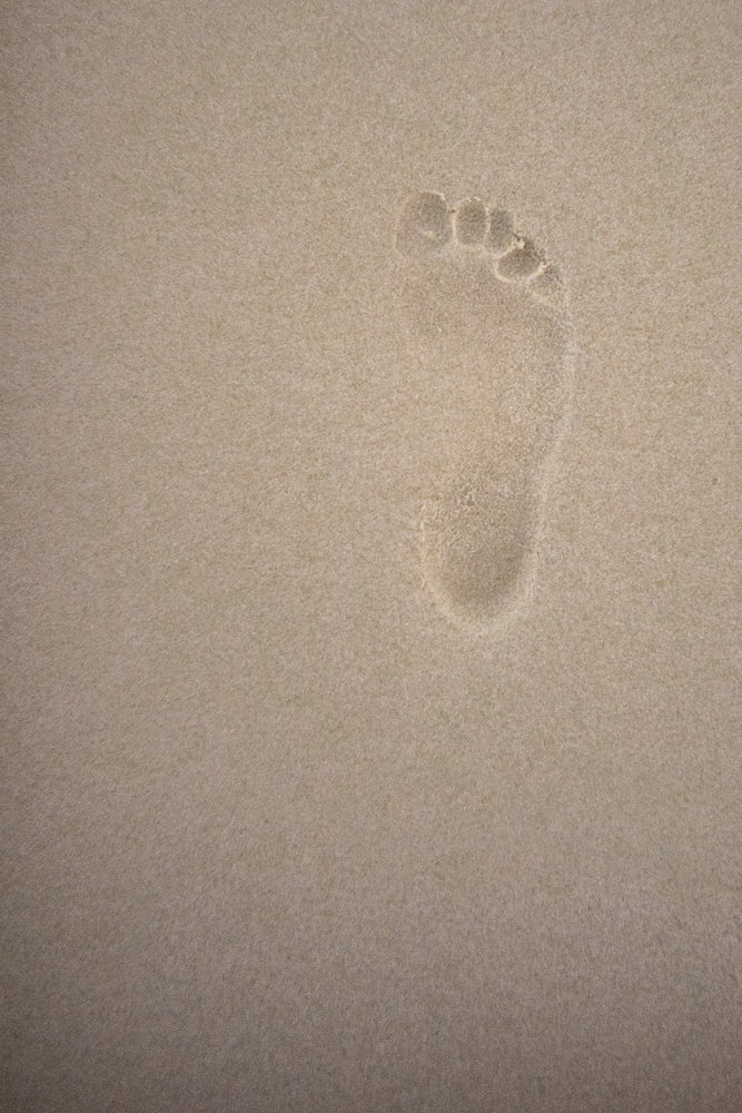 A single, clear footprint pressed into fine, light brown sand. The impression shows the shape of a bare foot, including the heel, arch, ball, and five toes.