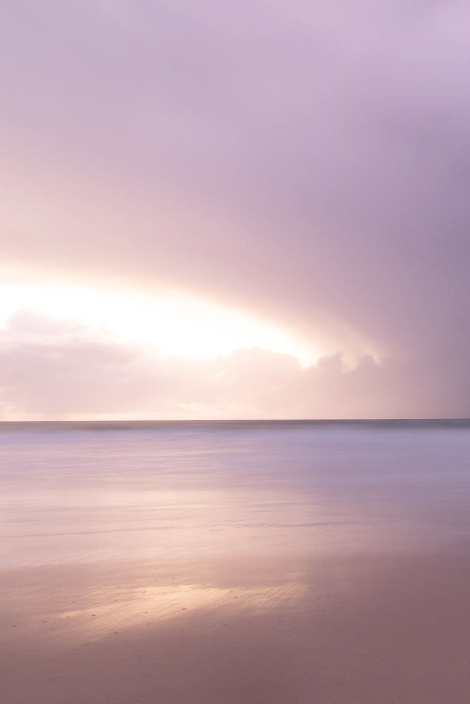 A serene, long-exposure shot of a beach at dusk. The sky is filled with soft purple clouds, with a bright golden light breaking through on the horizon. The ocean is a smooth, blurred expanse of lavender, reflecting the sky's colors. The wet sand in the foreground is also a muted purple, mirroring the gentle waves.