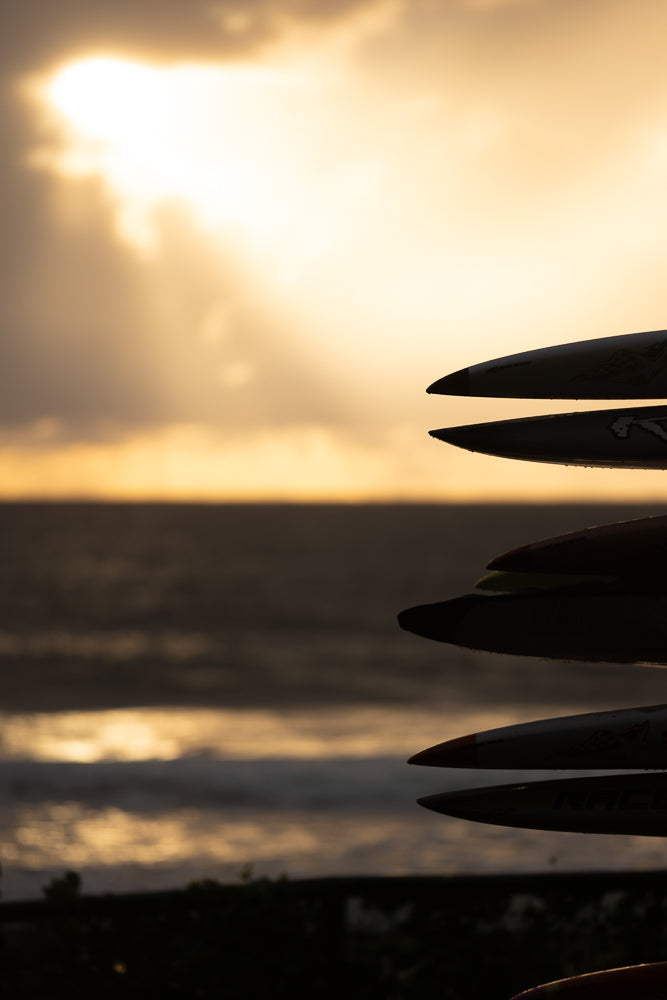 A stack of surfboards in silhouette against a golden sunset sky over the ocean. The tips of the boards are visible, with water droplets on them.