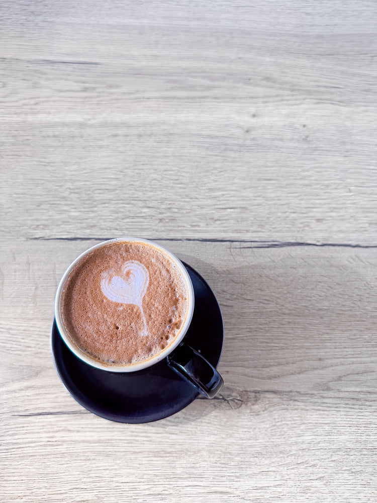A close-up, top-down view of a dark blue mug filled with a frothy, brown beverage, likely coffee or hot chocolate, with latte art in the shape of a heart on top. The mug sits on a matching dark blue saucer, both placed on a light-colored wooden surface.