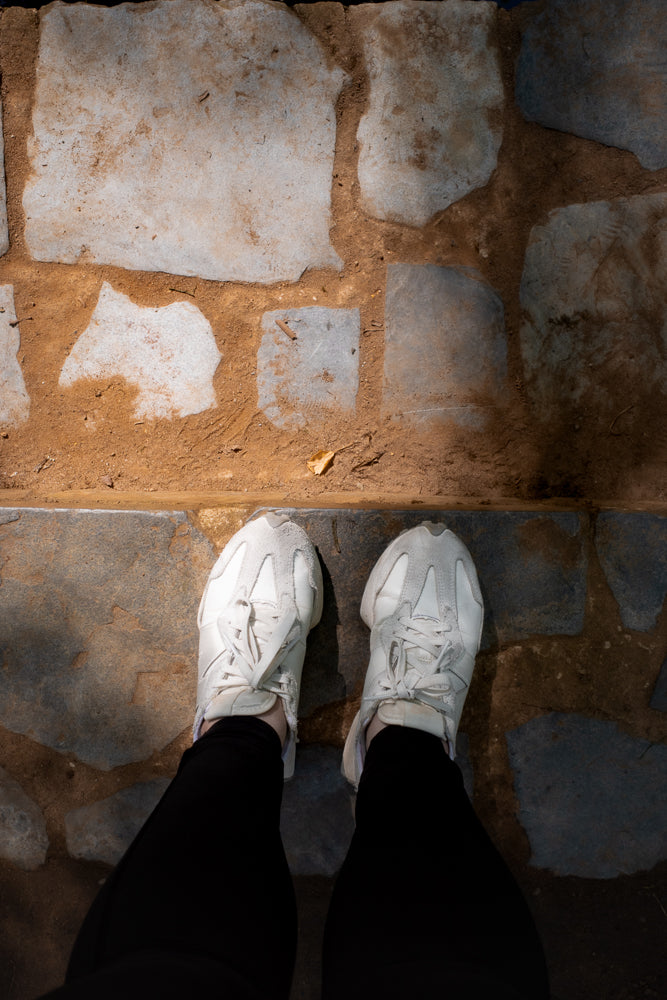 A top-down view of a person wearing white sneakers and black pants standing on a path made of irregular stone slabs and dirt.