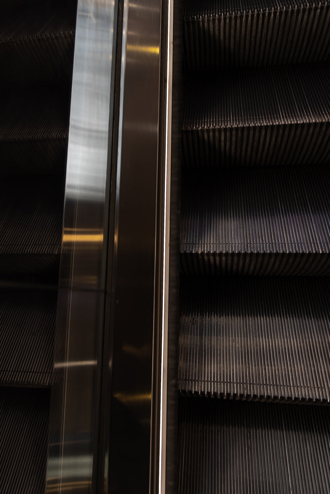 Close-up of an escalator with dark, ribbed steps and a polished metal side panel. The steps are angled, creating a sense of depth and movement.