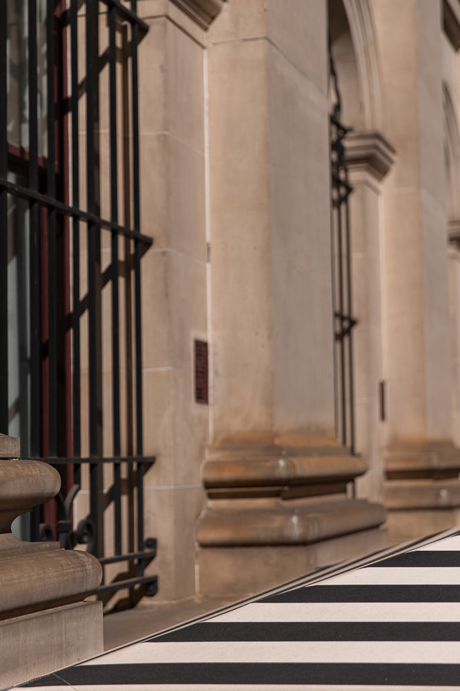 A close-up shot of a building facade with large stone columns and arched windows. A black metal railing is visible in the foreground, casting shadows on the building. The lower part of the image features a black and white striped pattern.