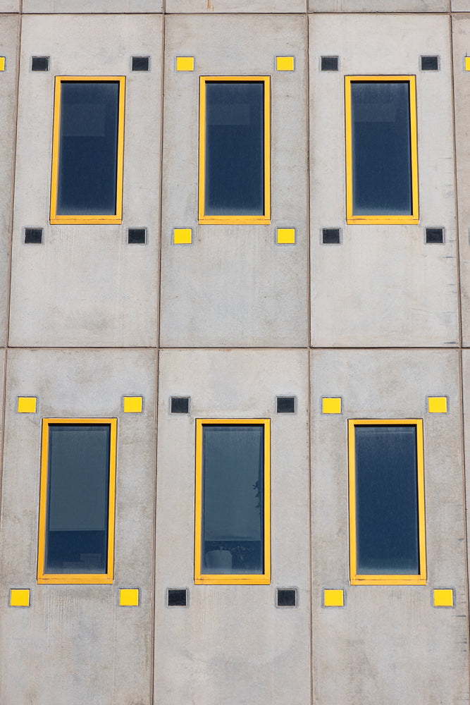 A close-up, straight-on view of a concrete building facade with a repeating pattern of six rectangular windows. The windows have dark blue glass and bright yellow frames. Each window is flanked by two small square accents, alternating between black and yellow.