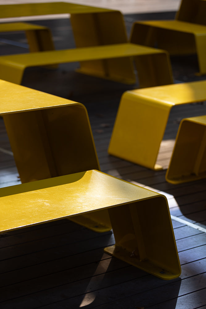Several bright yellow, modern benches are arranged on a dark wooden deck. Sunlight casts dramatic shadows across the scene, highlighting the curved edges of the benches.