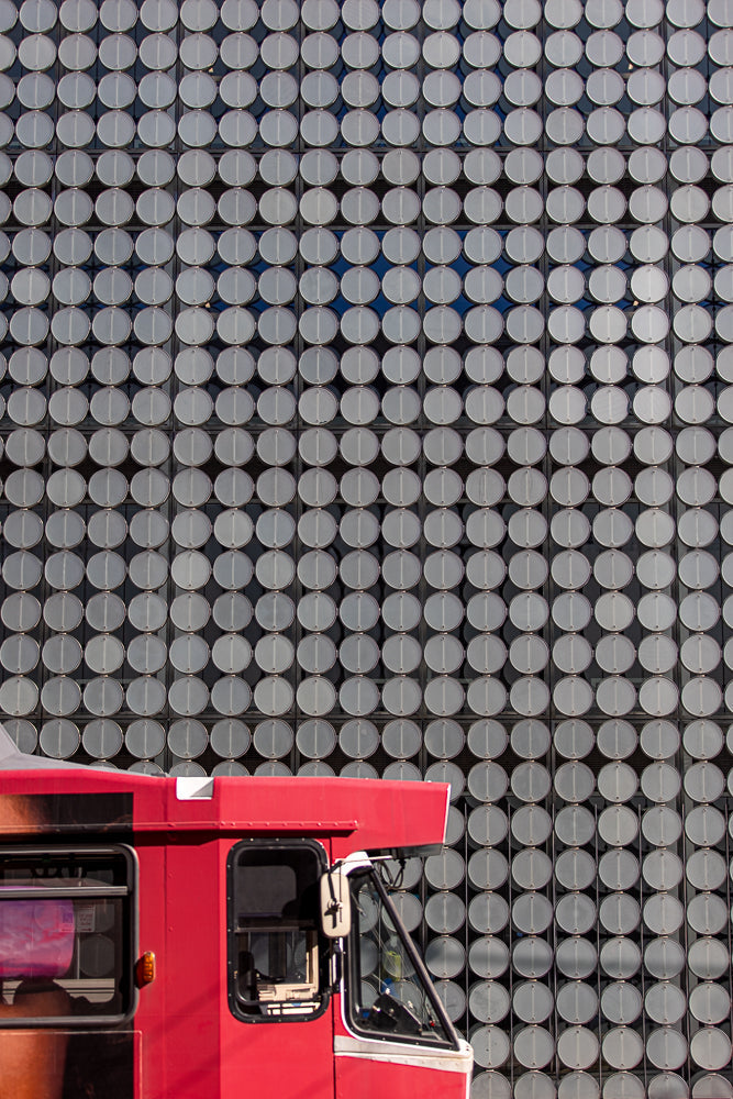 A red bus is parked in front of a building with a facade covered in a grid of circular, metallic panels. The panels have a matte silver finish and are arranged in neat rows and columns, creating a textured, repetitive pattern.