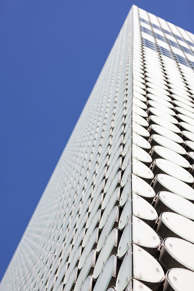 A modern building with a white, textured facade made of oval panels, set against a clear blue sky. The perspective is from a low angle, emphasizing the building's height and unique design.
