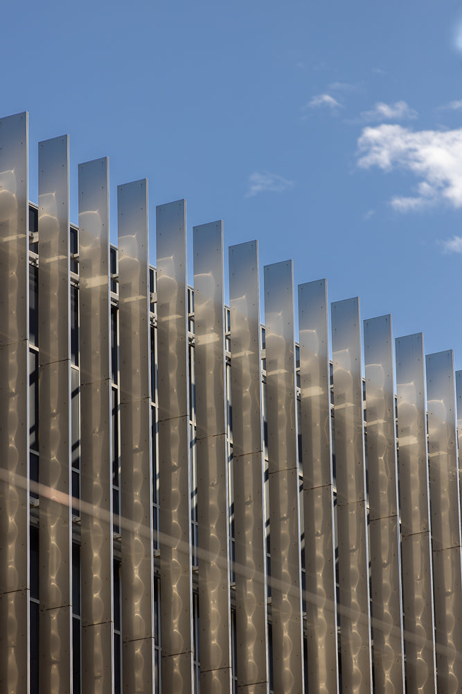 A modern building facade with vertical metallic fins reflecting sunlight and clouds against a bright blue sky.
