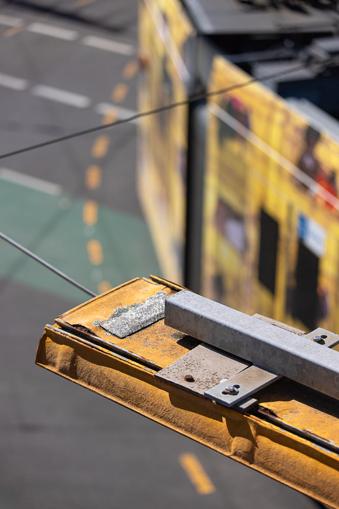 Close-up of a weathered yellow metal surface with a silver-colored metal bracket attached by bolts. In the blurred background, a yellow tram with graphics is visible, along with a street with green and white markings.