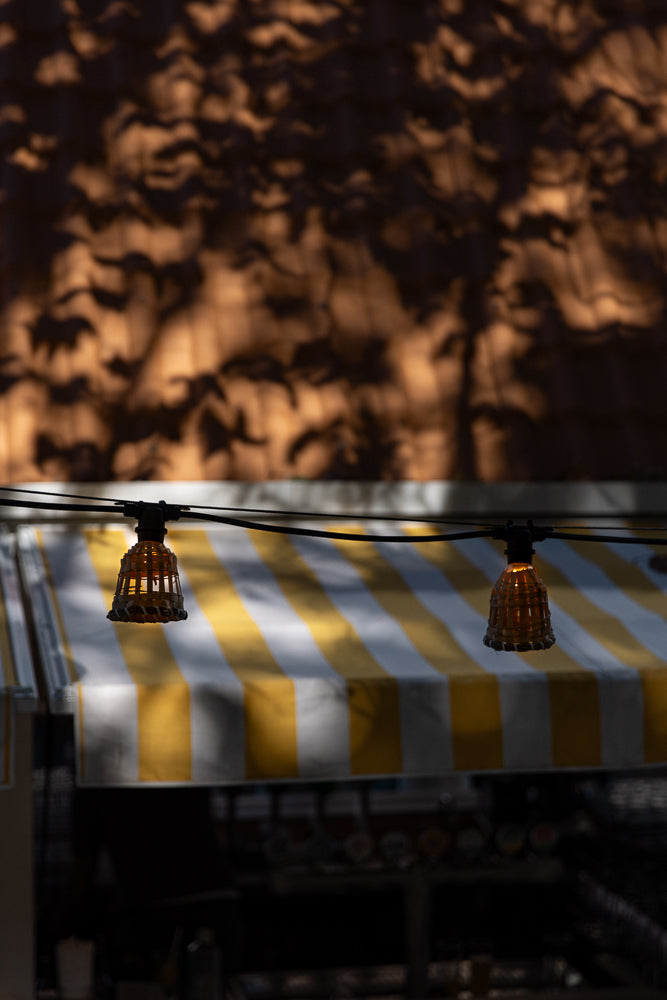 A string of lights with woven bamboo shades hangs above a yellow and white striped awning. The background is a wall with dappled shadows of leaves.