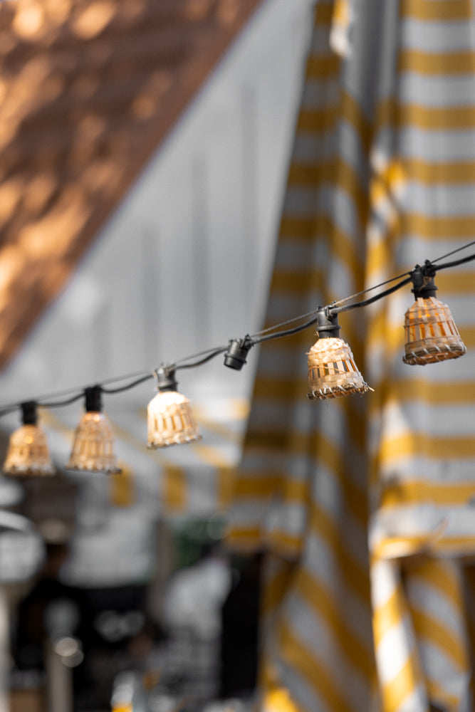 A string of lights with woven shades hangs in front of blurred yellow and white striped awnings and a brown textured surface.