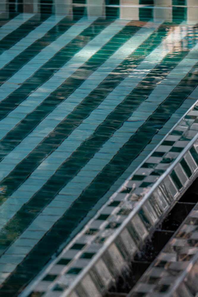 Diagonal stripes of dark green and light blue tiles are visible under rippling water. The tiles appear to be submerged in a pool or fountain, with light reflecting off the surface.