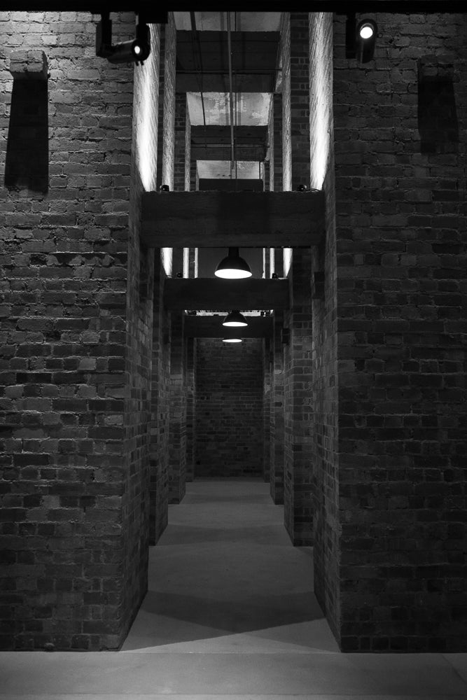 A black and white image of a long, narrow hallway with brick walls. The hallway recedes into the distance, with multiple openings and beams creating a sense of depth. Lights are visible along the ceiling and on the beams, illuminating the space.