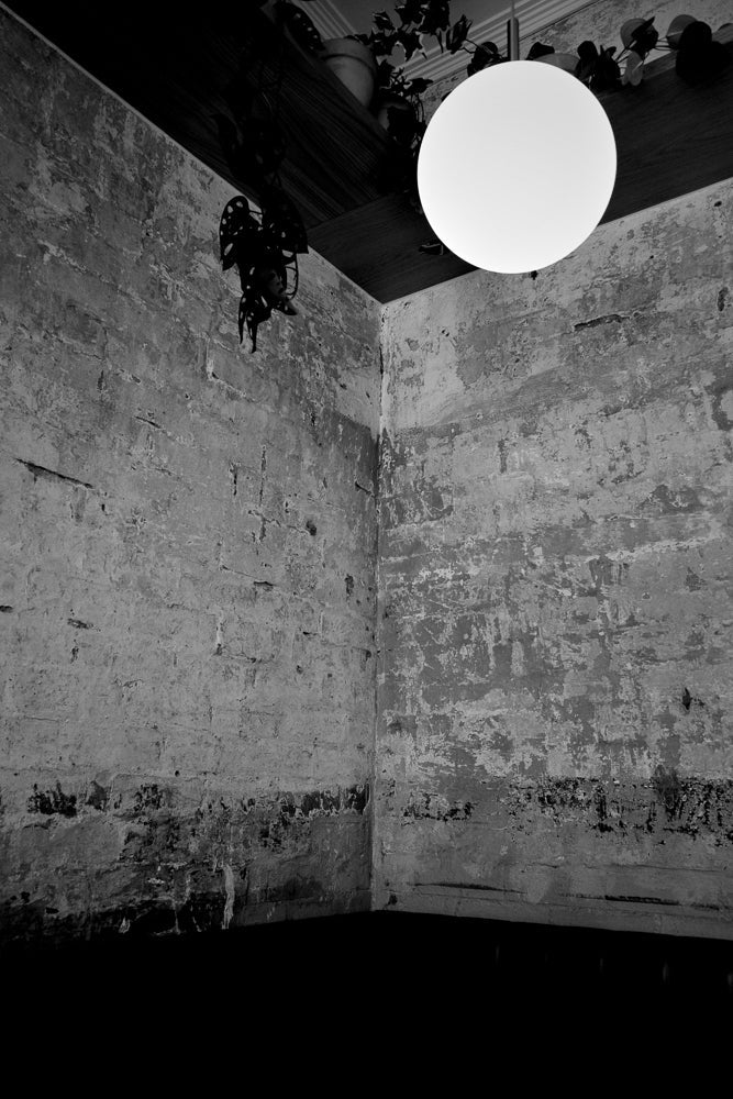 A black and white image of a dimly lit corner with rough, textured walls. A large, round, white light fixture hangs from the ceiling, illuminating the space.