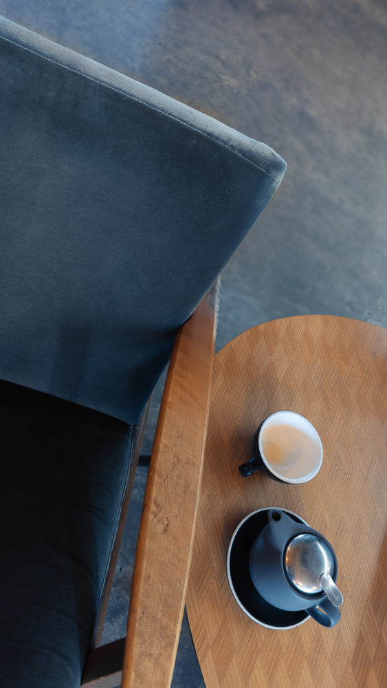 A close-up, overhead view of a dark blue armchair next to a round wooden table. On the table sits a white cup with a light brown beverage and a dark blue teapot with a silver lid.